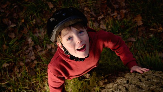 Child in a red sweater and black helmet looks up from the ground among fallen leaves.