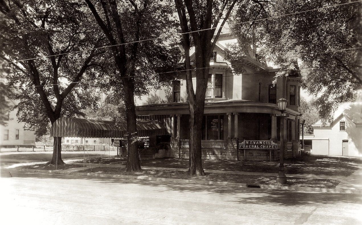 Sepia-toned historic house with wraparound porch beneath large trees on a quiet street.