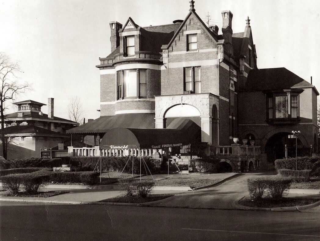 Historic brick mansion with a rounded porch and ornate turrets beside a street.