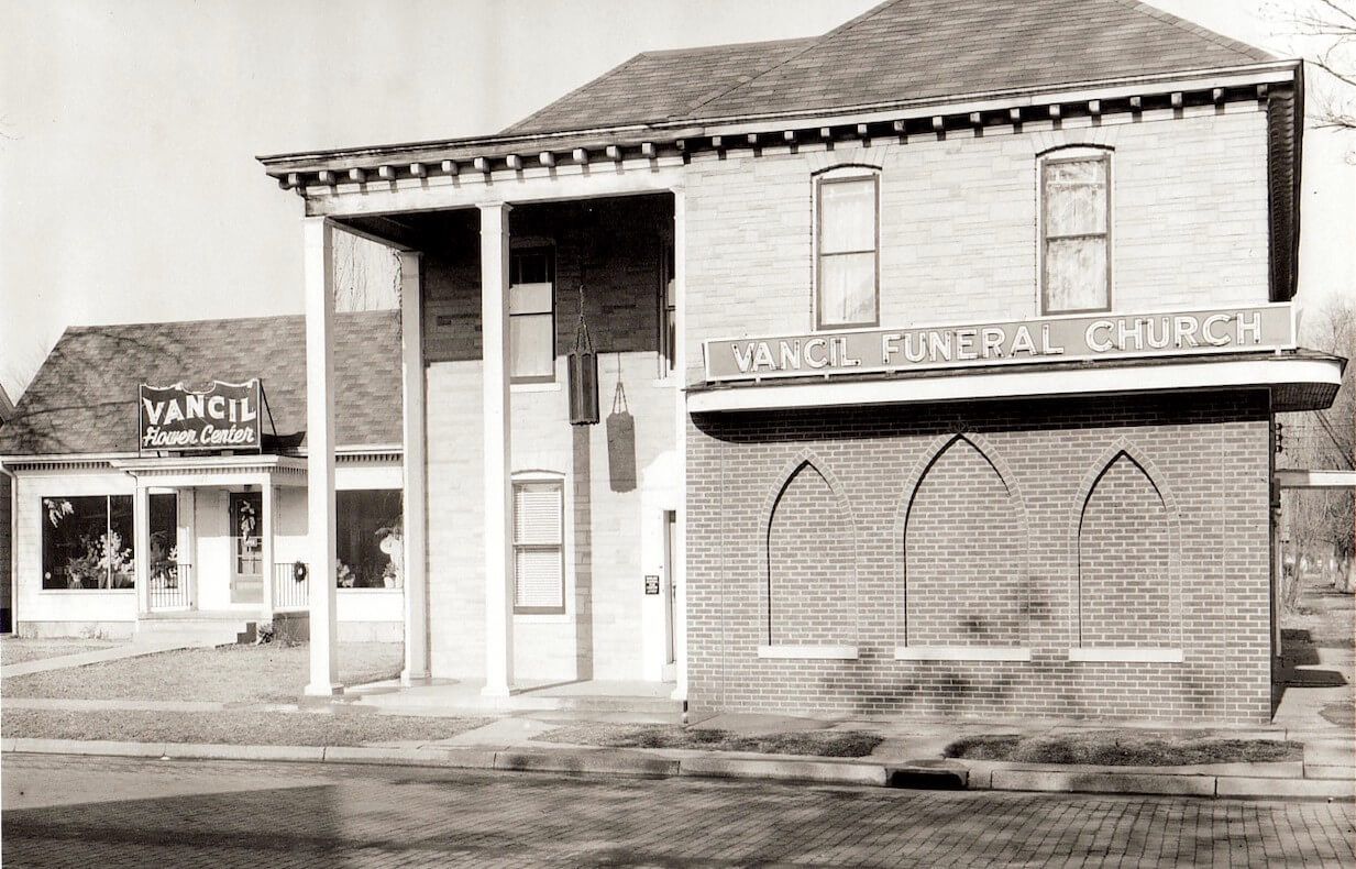 Black-and-white photo of a brick building with a columned entrance and adjacent storefronts