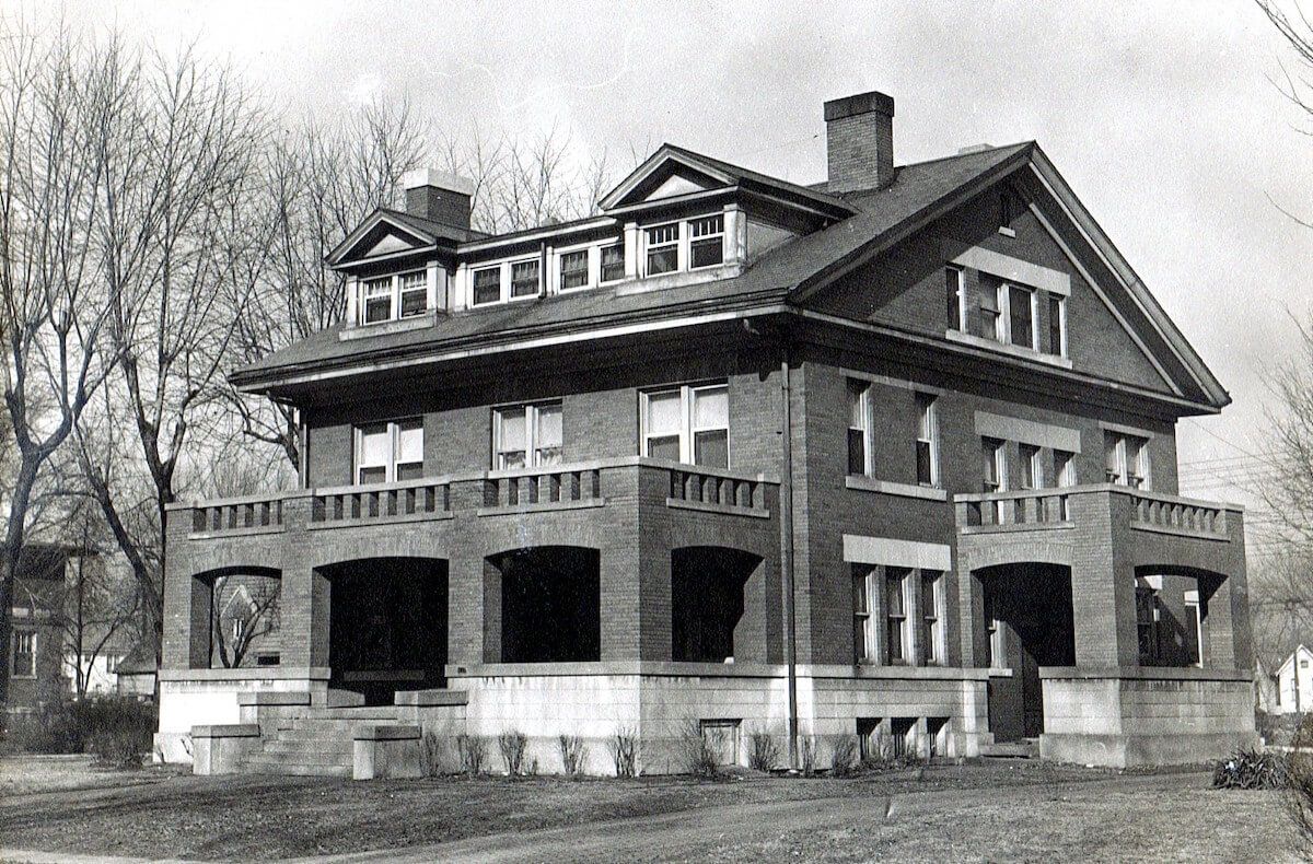 Black-and-white photo of a large historic house with a wraparound porch and dormer windows