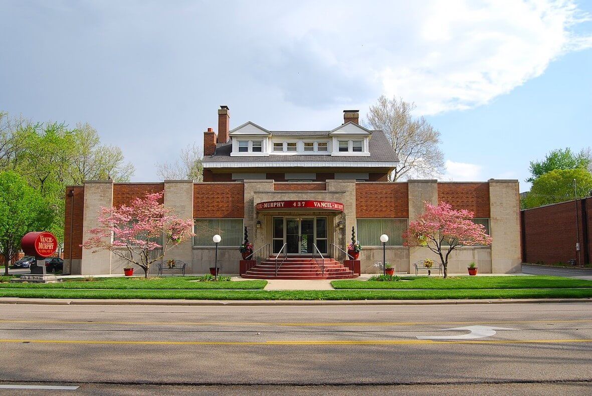 A brick building with a red entrance, decorative trees, and a street in front.