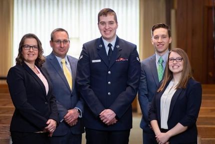 Five people in business and military attire pose together indoors against a wood-paneled background.