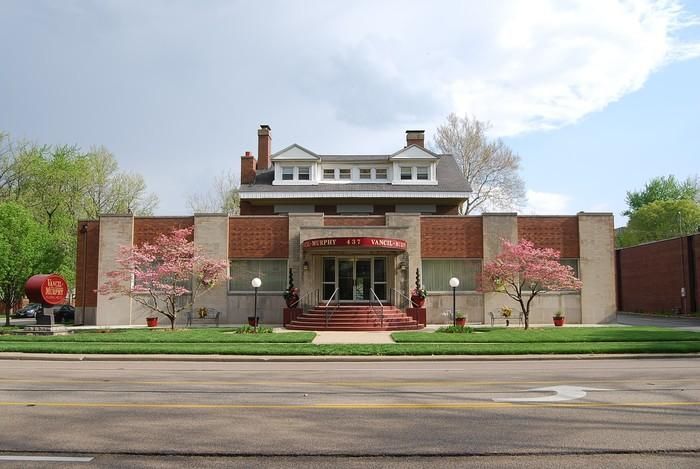 Front view of a brick building with a red entrance, trees, and a street in the foreground