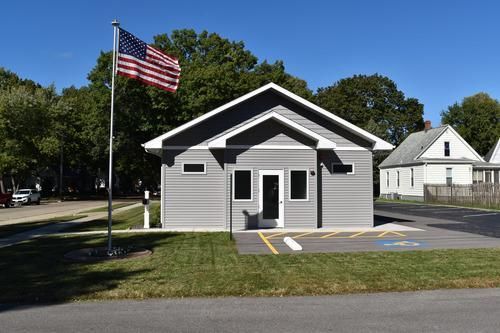Small gray building with an American flag, front entrance, and parking lot on a sunny day