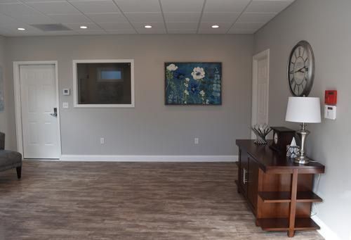 Empty lobby with gray walls, wood floor, framed art, a side table, lamp, and wall clock.