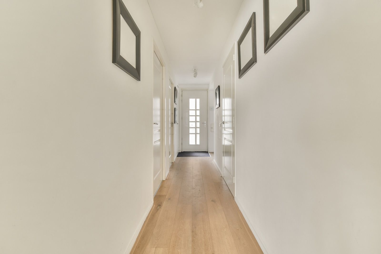 Narrow hallway with white walls, light wood floor, framed art, and a door at the end.