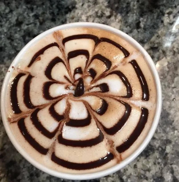 A cappuccino with chocolate syrup swirled into a flower pattern on foamy milk.