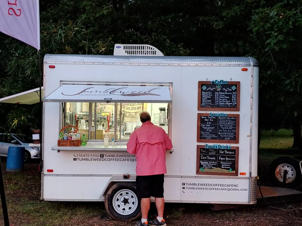 Food truck with service window and menu boards, a person in a pink jacket ordering at dusk.