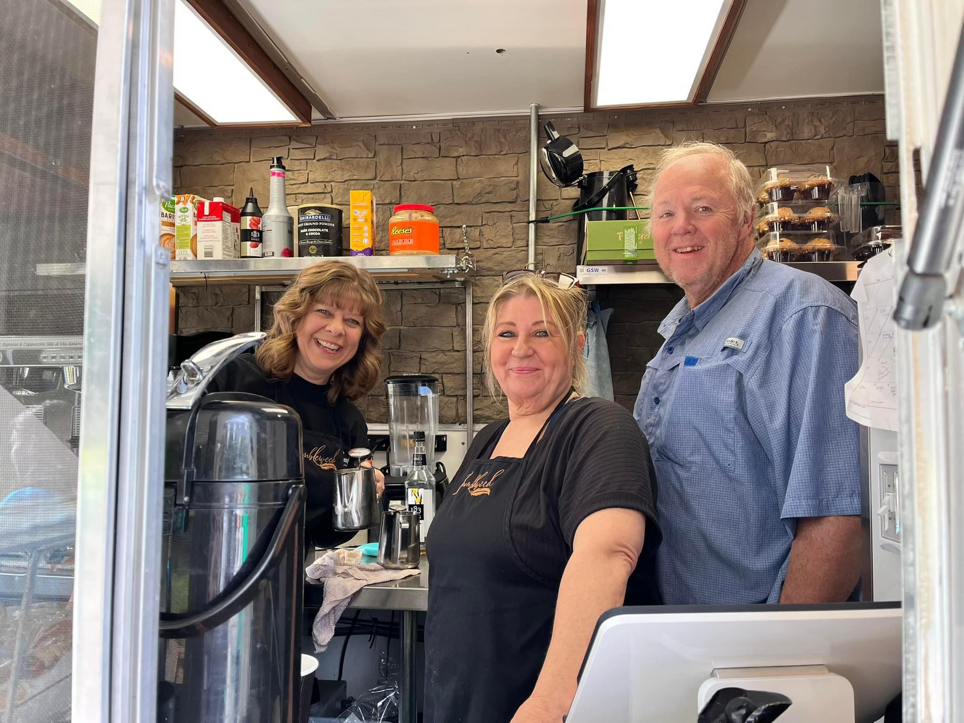 Three people smiling in a kitchen, standing behind a counter with appliances and shelves in the background.