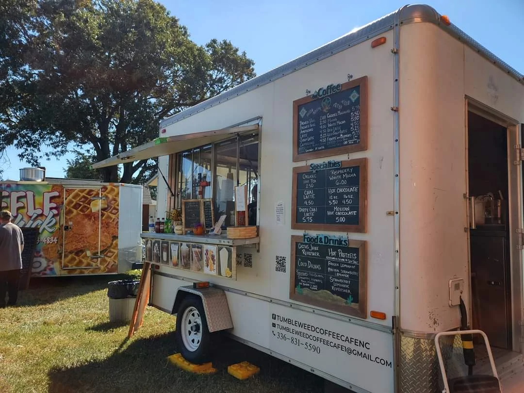 White food trailer with menu boards and service window at an outdoor fair, with another booth beside it