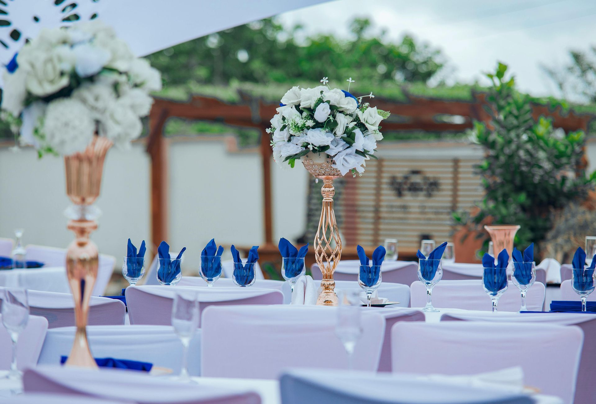 Elegant outdoor banquet table with white linens, blue napkins, and tall white floral centerpieces under a gazebo