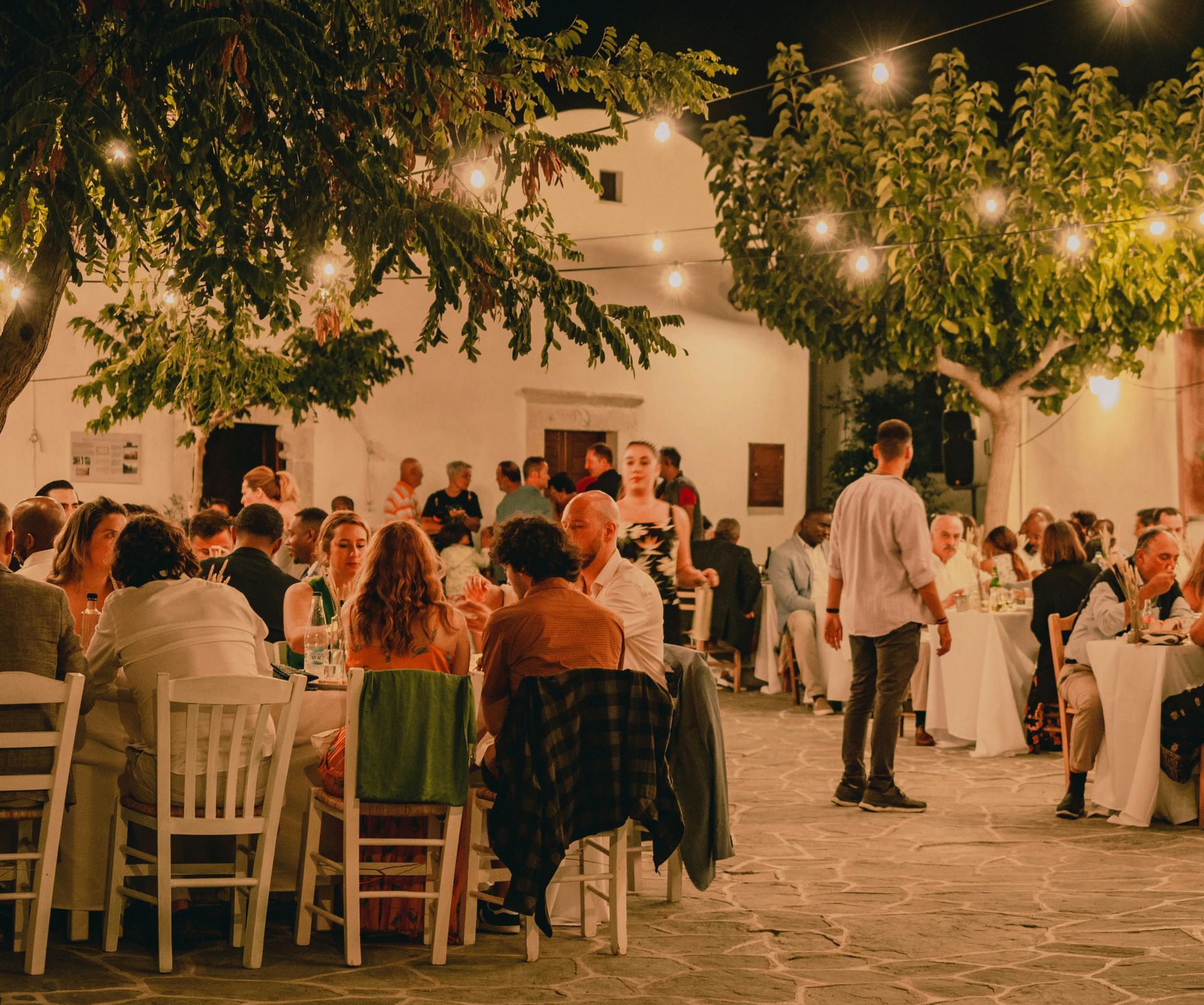 Outdoor evening gathering with seated guests under string lights in a courtyard, with a standing speaker nearby