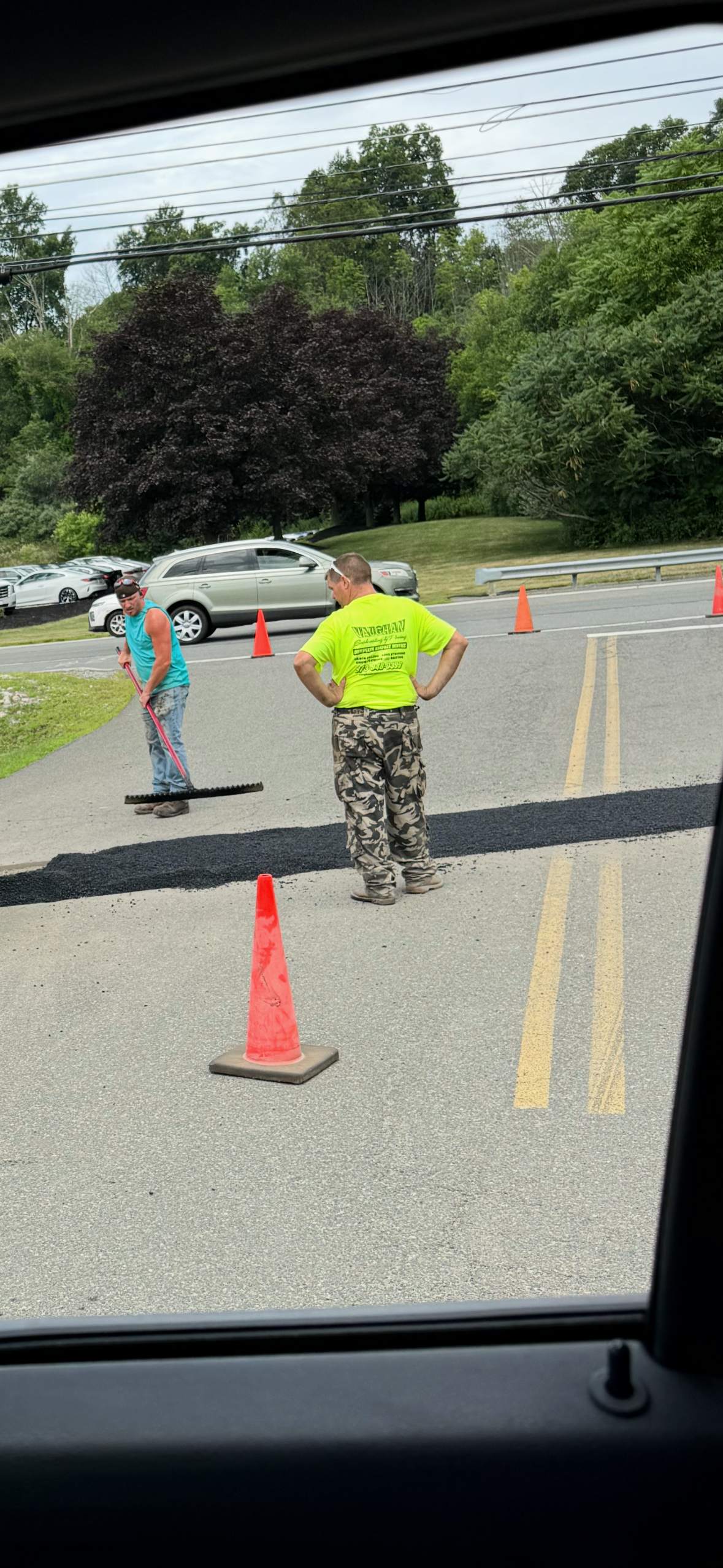 Workers in high-visibility orange gear use rakes to spread fresh asphalt on a sunny road near a manhole cover.