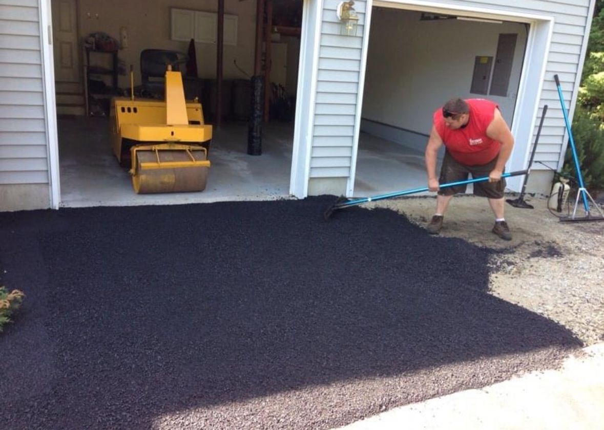 A person in work boots uses a shovel to fill a pothole with fresh, black asphalt.