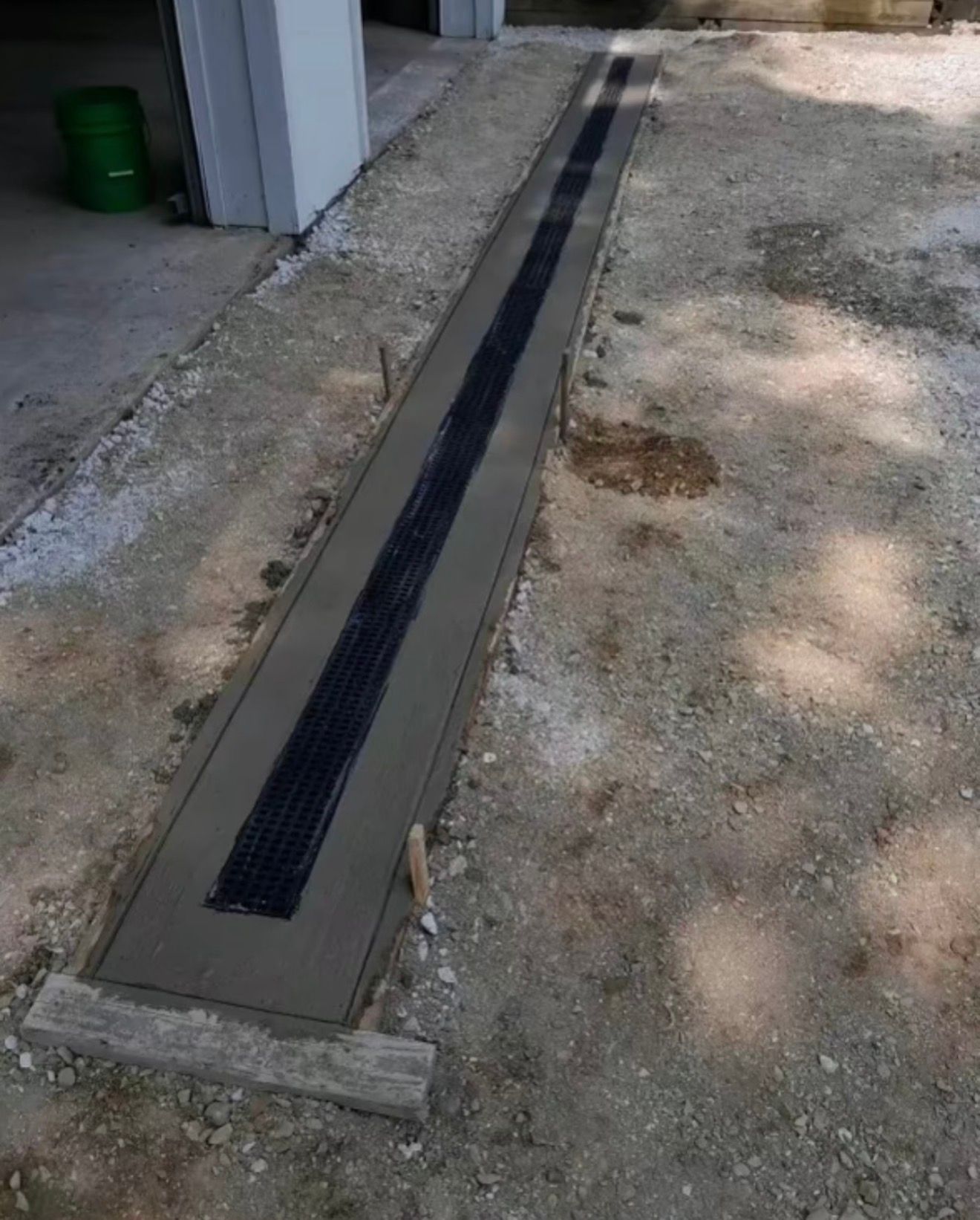 A newly installed black trench drain set in a concrete strip alongside a gravel driveway.