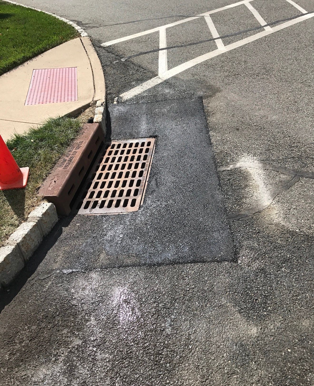 A rectangular storm drain grate set into a patch of fresh black asphalt at the edge of a curb and paved street.
