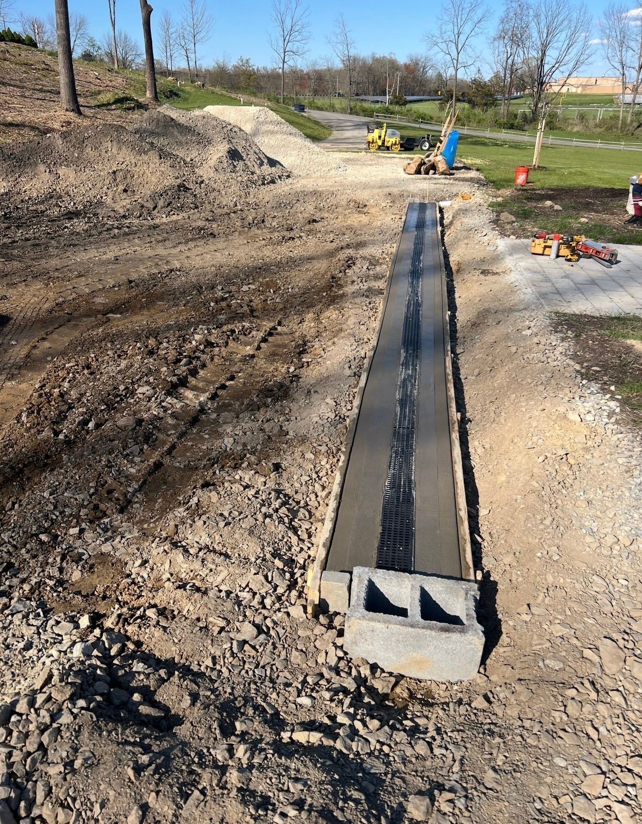 A construction site showing a fresh concrete footing with a drainage channel and a single concrete block at the end.