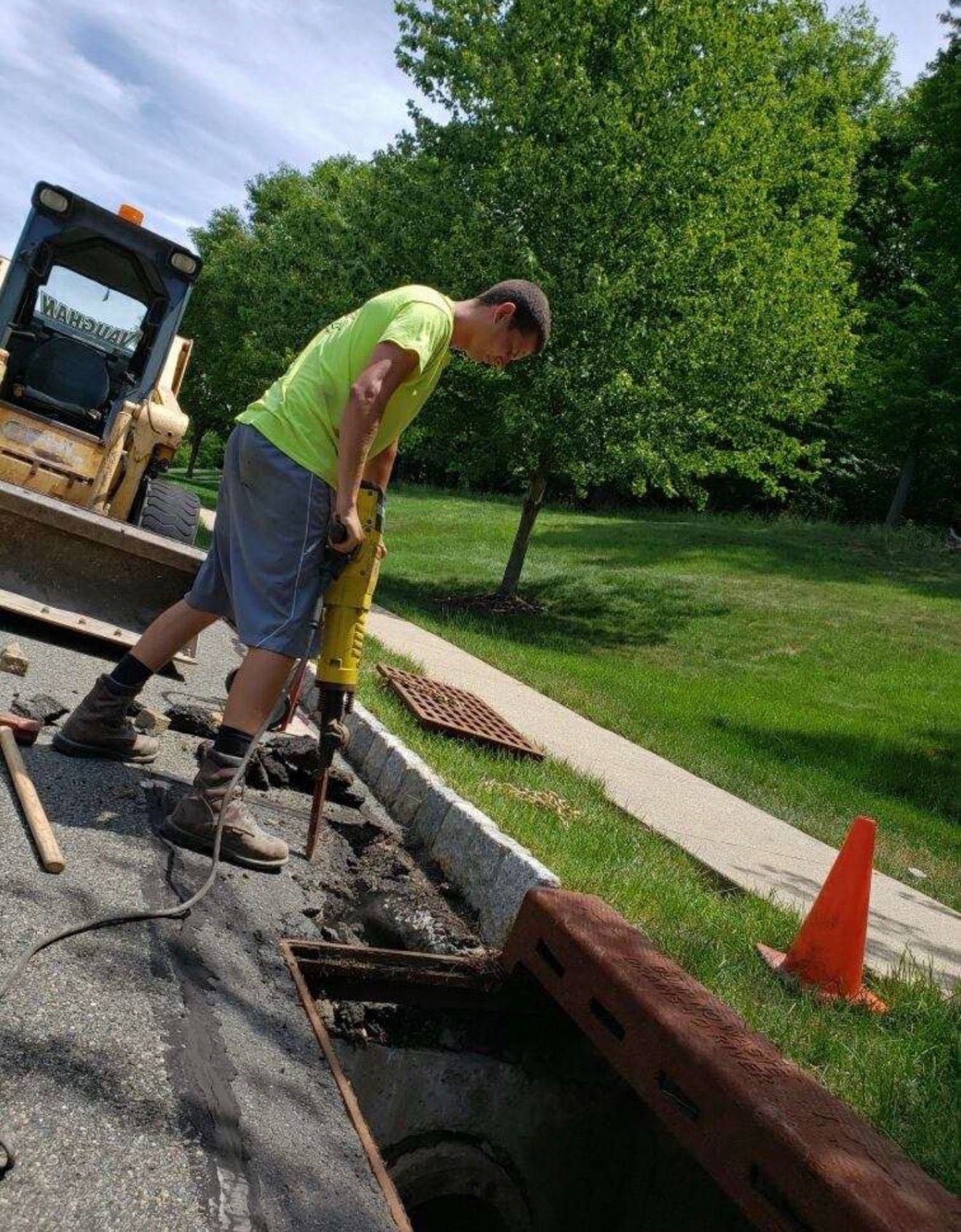 A person in a bright yellow shirt uses a jackhammer to break up asphalt next to a construction vehicle and a safety cone.