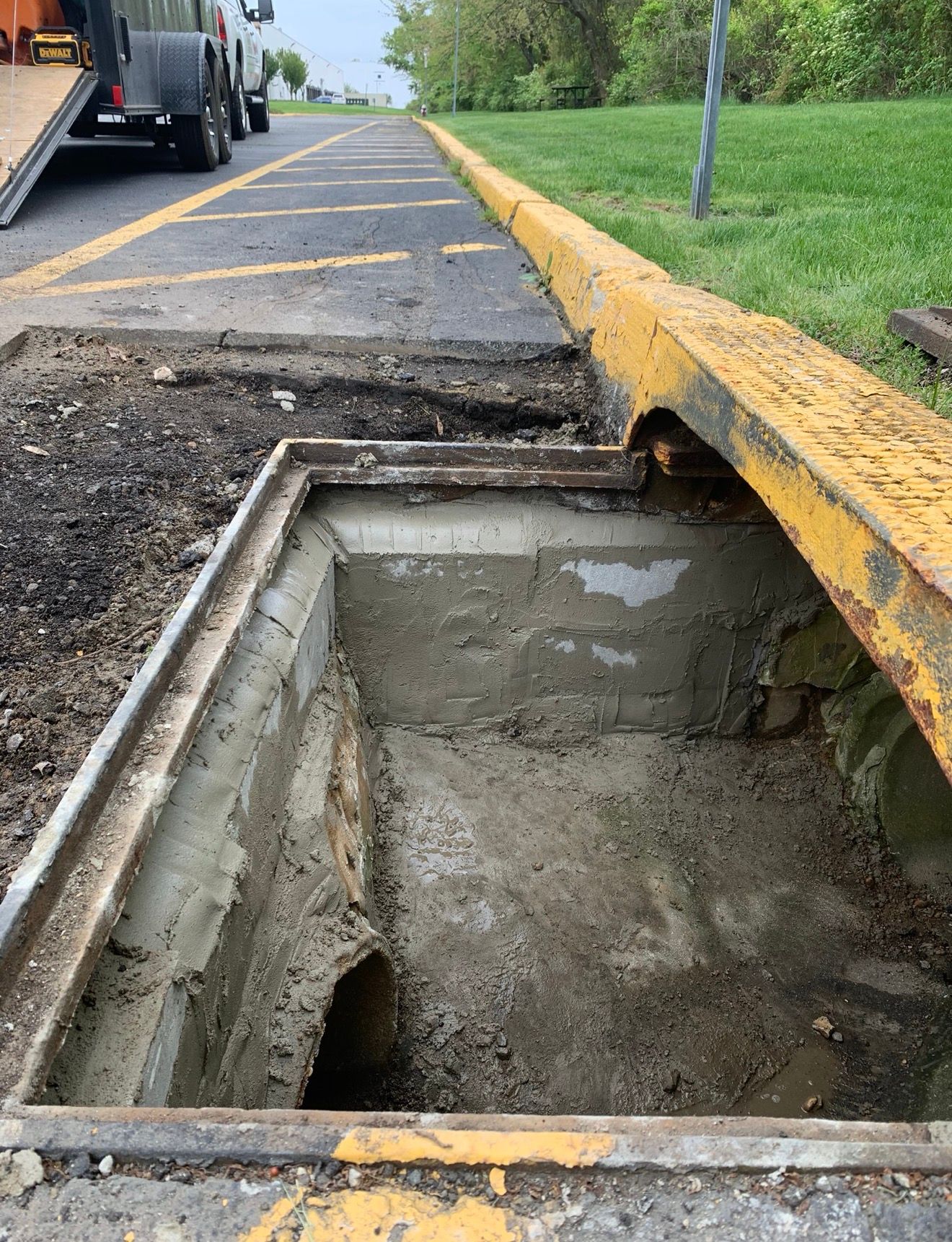 An open concrete storm drain box sits along a yellow curb near a road with a trailer parked in the background.