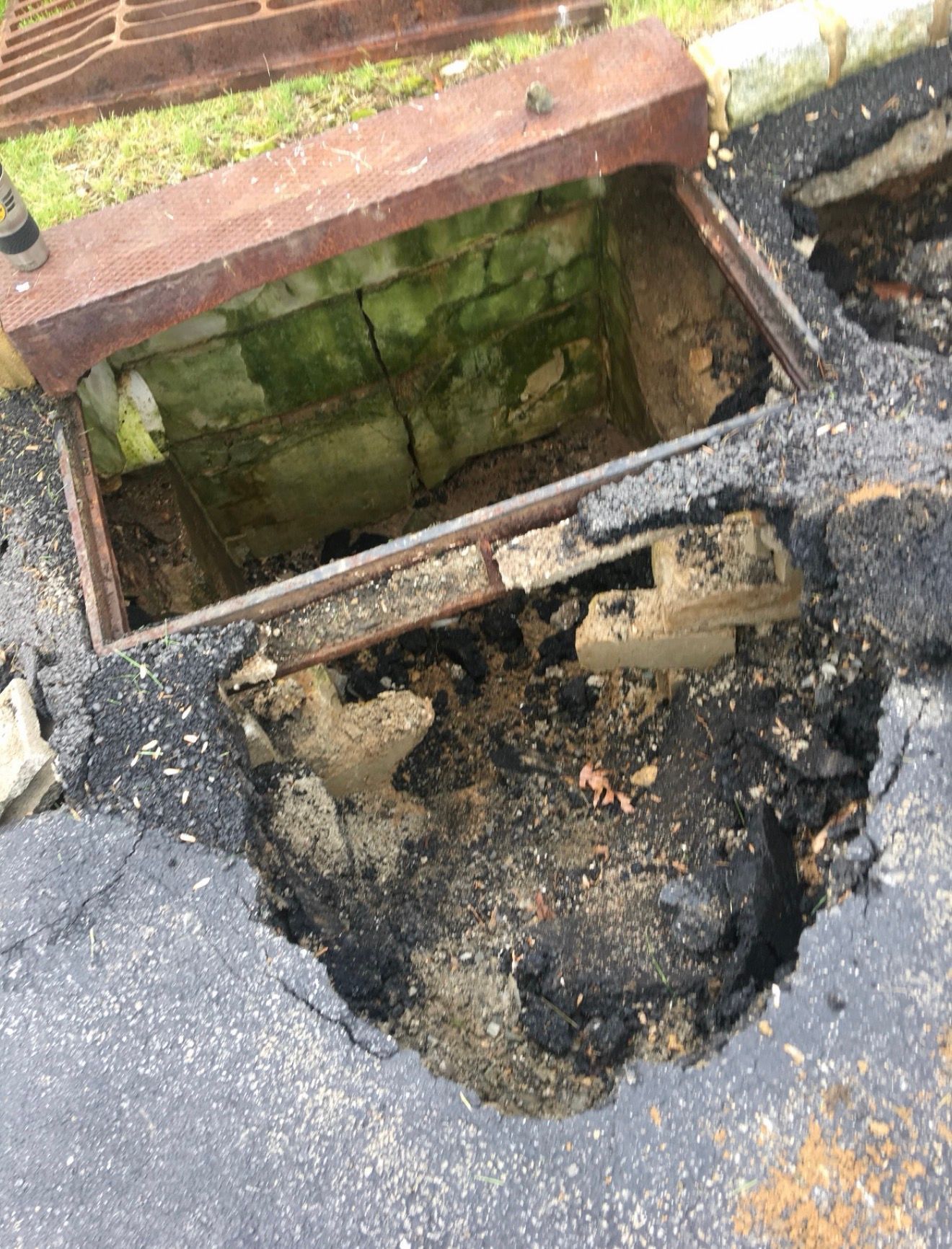A damaged asphalt curb featuring a large, crumbling hole next to a deep, rectangular drainage inlet with mossy walls.
