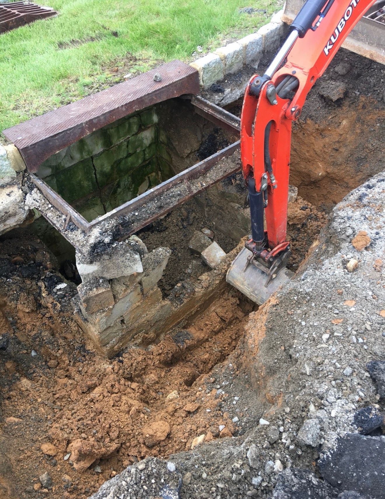 An orange excavator digging in the dirt next to an open, brick-lined underground utility or drainage structure.