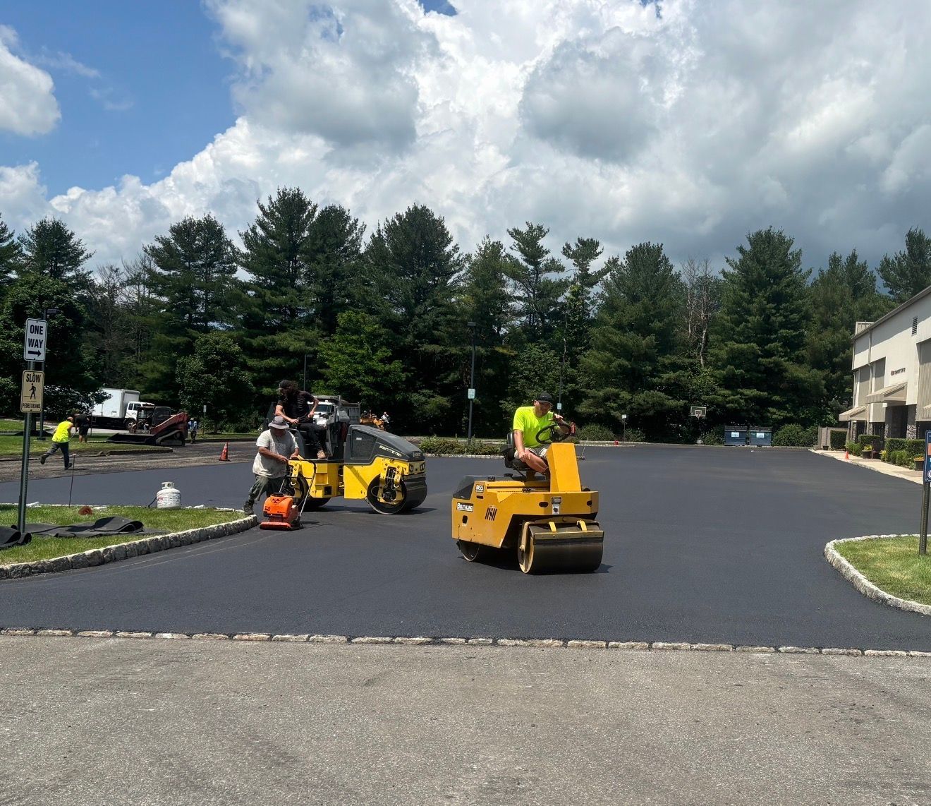 Workers operate two yellow asphalt rollers on a freshly paved black parking lot under a cloudy blue sky.