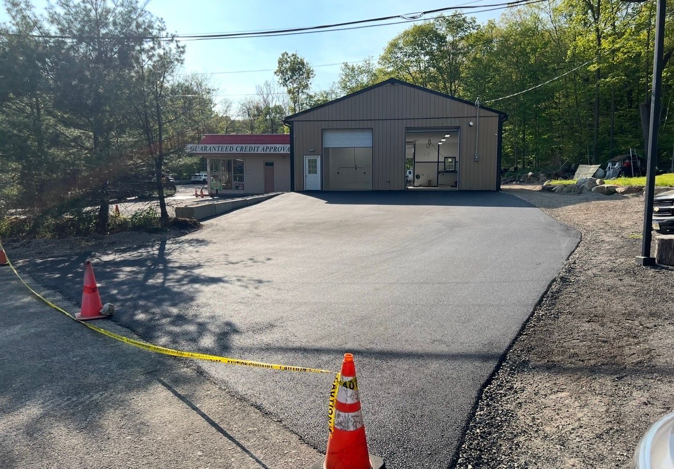 A newly paved asphalt driveway leads to a metal-sided workshop building under a bright sky with orange traffic cones.