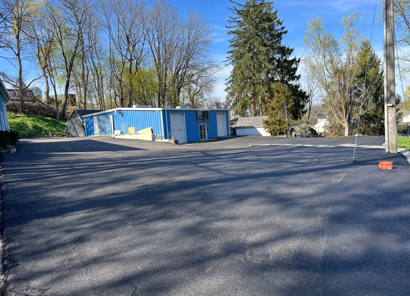 A large, freshly paved asphalt parking lot leads to a blue commercial building surrounded by trees under a clear blue sky.
