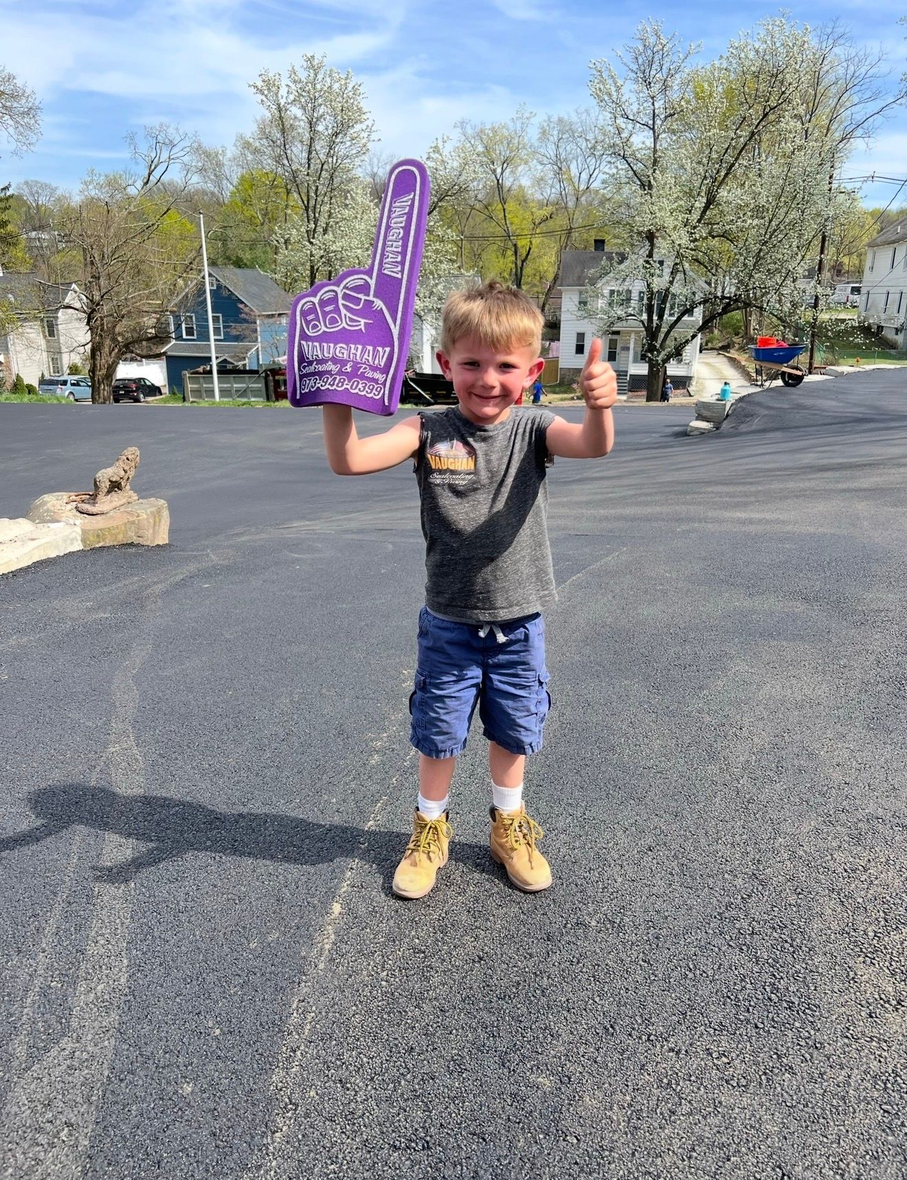 A young child standing on asphalt, smiling and giving a thumbs-up while holding a purple foam finger.