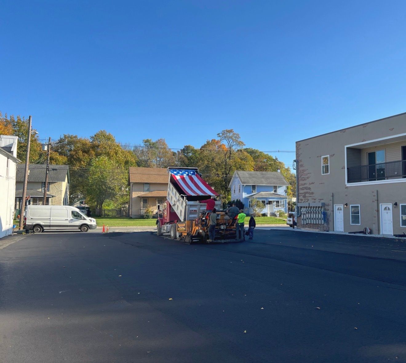 A construction crew operates a paving machine with an American flag-patterned topper in a paved lot near residential houses.