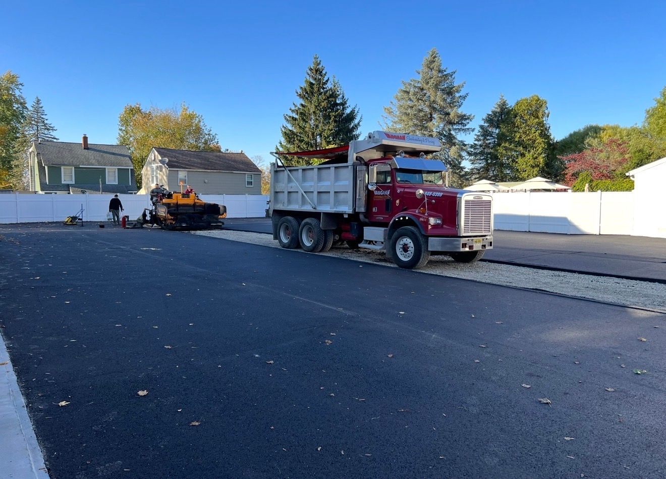 A red dump truck sits on a newly paved black asphalt lot next to construction equipment, with houses in the background.