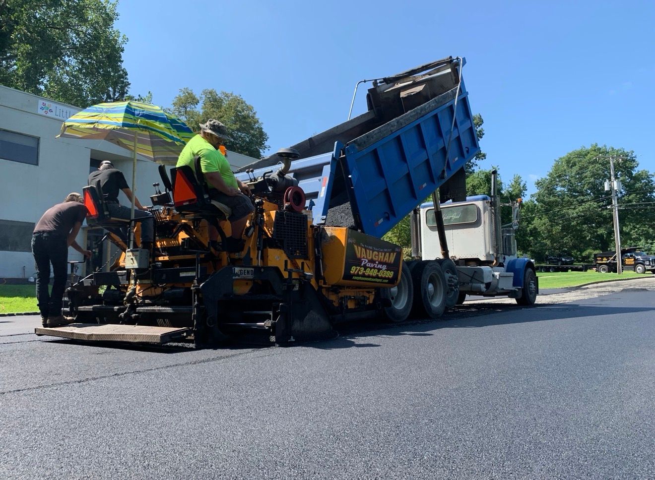 A dump truck pours asphalt into a yellow paving machine on a sunny day as workers operate the equipment on a paved road.