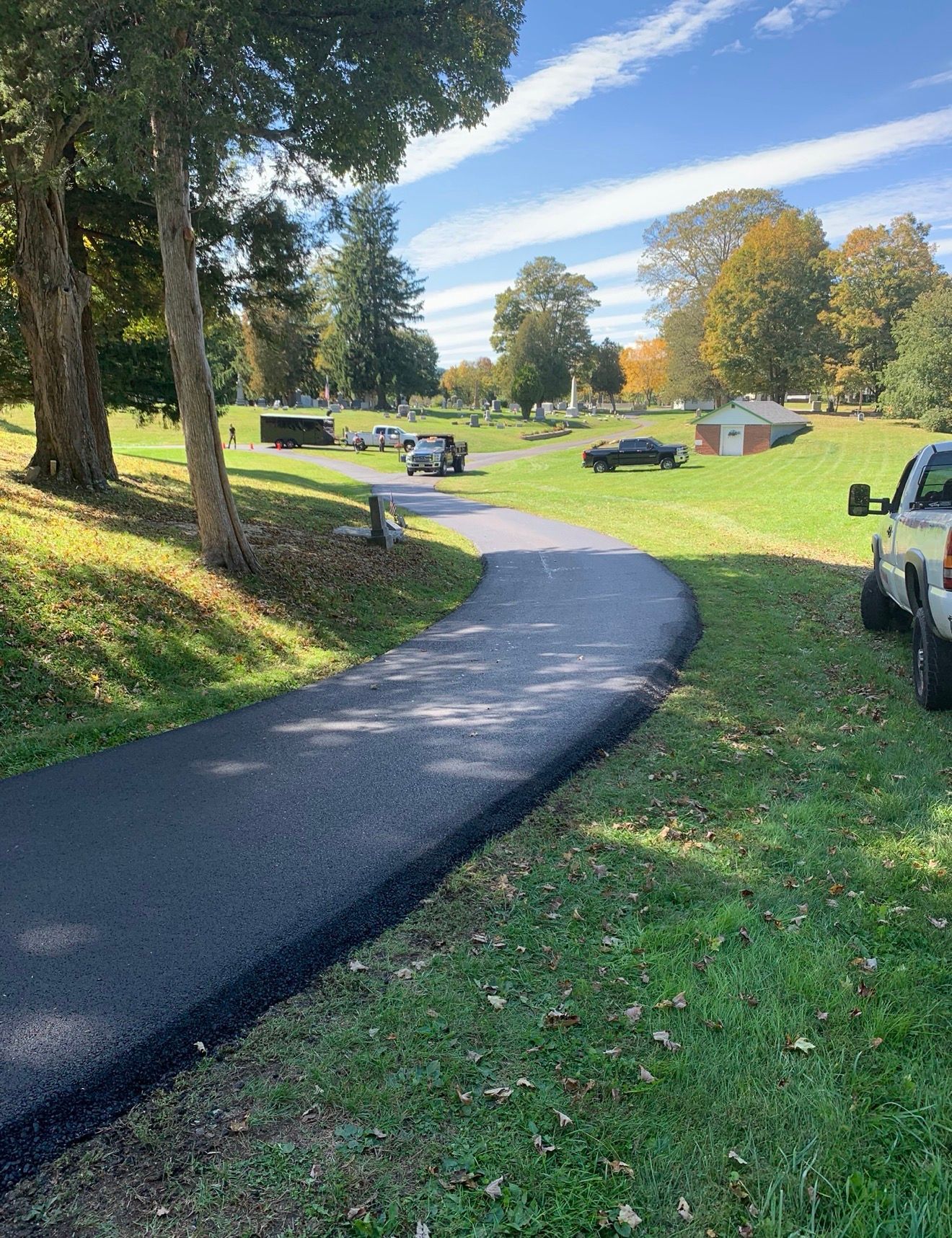 A freshly paved asphalt path winds through a grassy park with trees, several parked vehicles, and a small building.