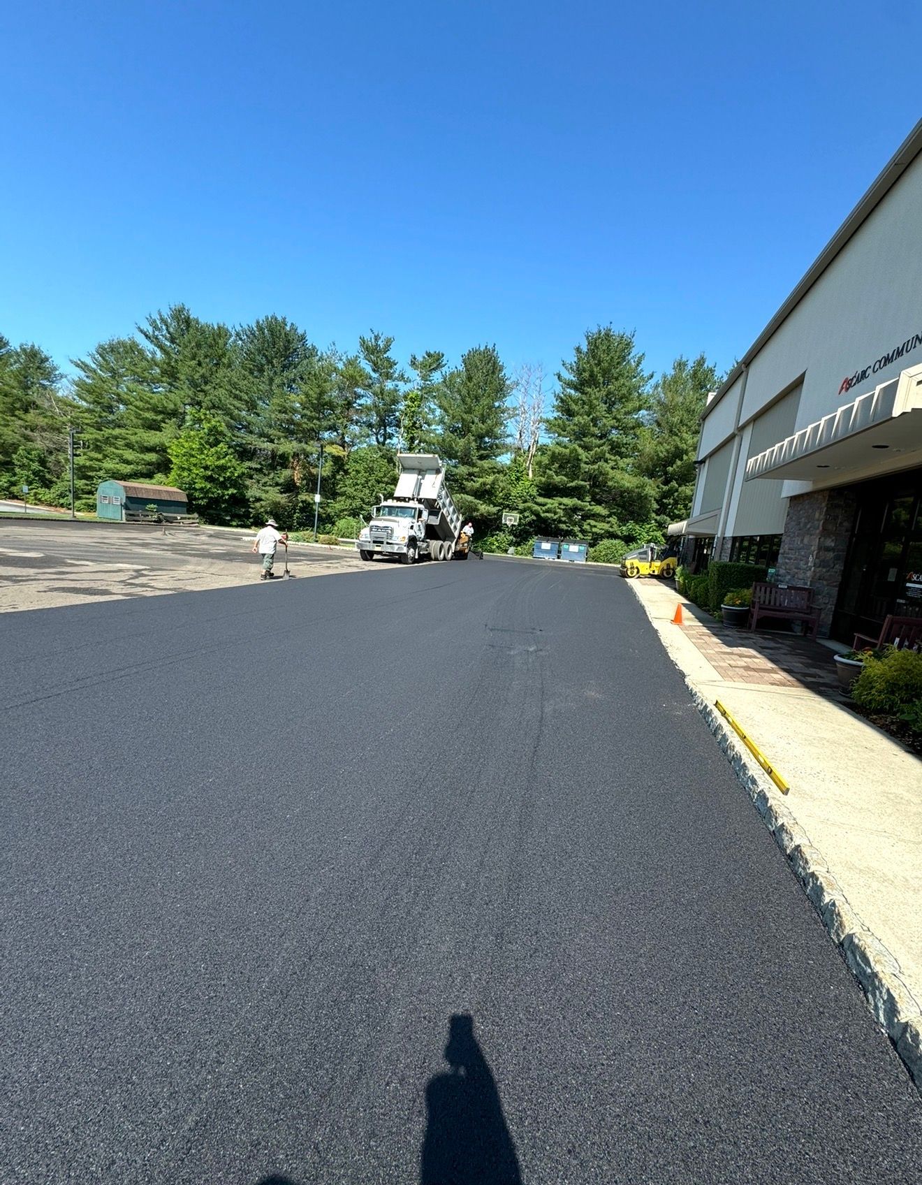 Freshly paved asphalt driveway leading to a building, with a work truck in the distance under a clear blue sky.