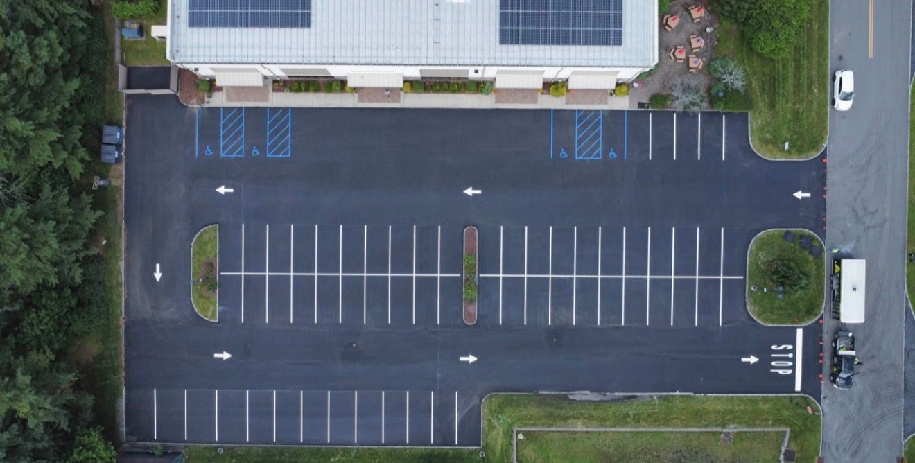 An aerial view of a paved parking lot next to a building with solar panels, showing parking stalls and painted traffic lanes.