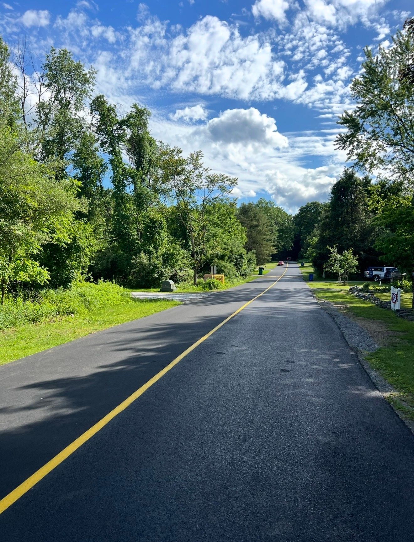A paved multi-use trail with a yellow center line winds through a lush, green wooded area under a partly cloudy blue sky.