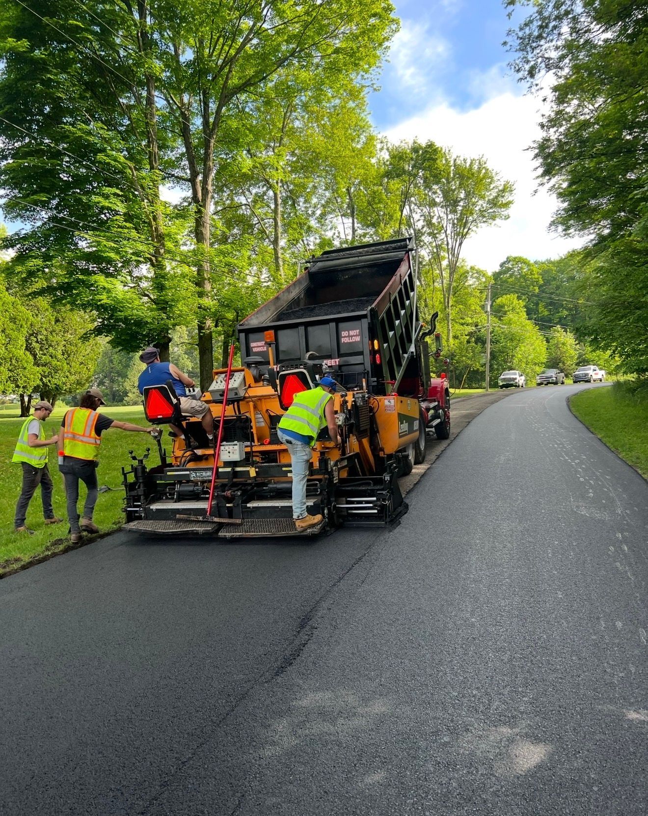Workers in neon vests operate a large orange paving machine applying fresh asphalt to a road lined with green trees.