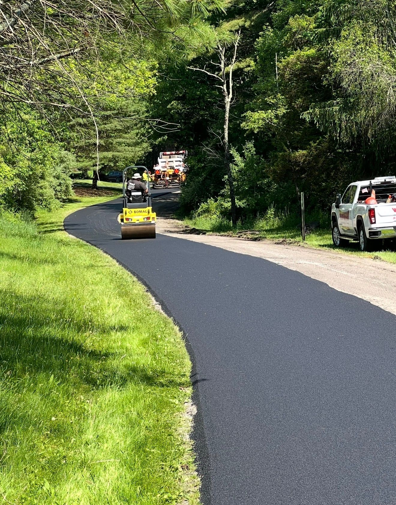 A yellow construction roller travels along a newly paved black asphalt path through a green, wooded area.