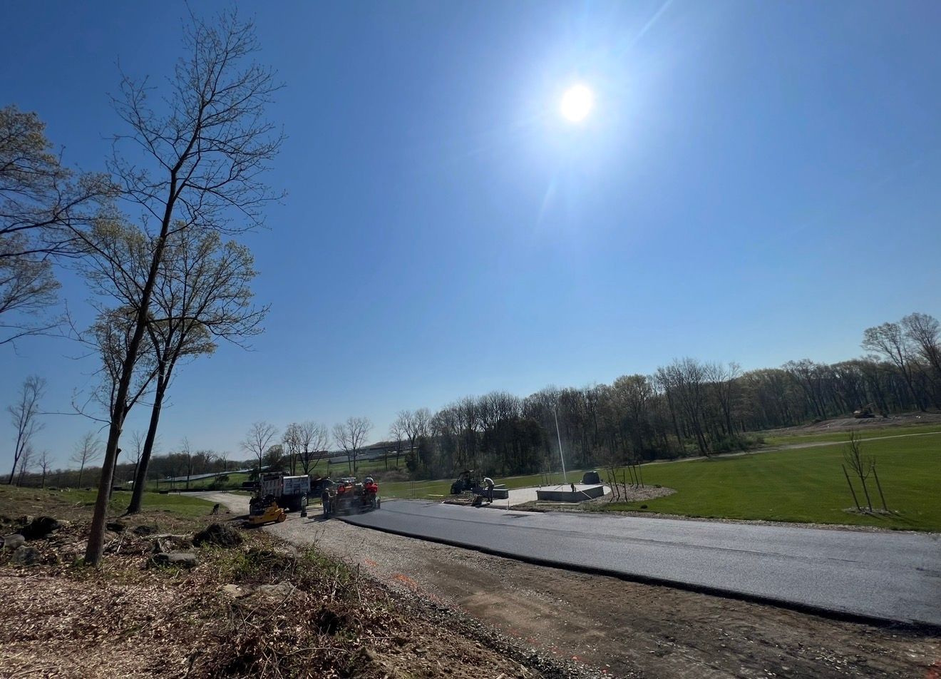 Construction workers pave a new asphalt road under a bright, sunny sky near a grassy field and trees.