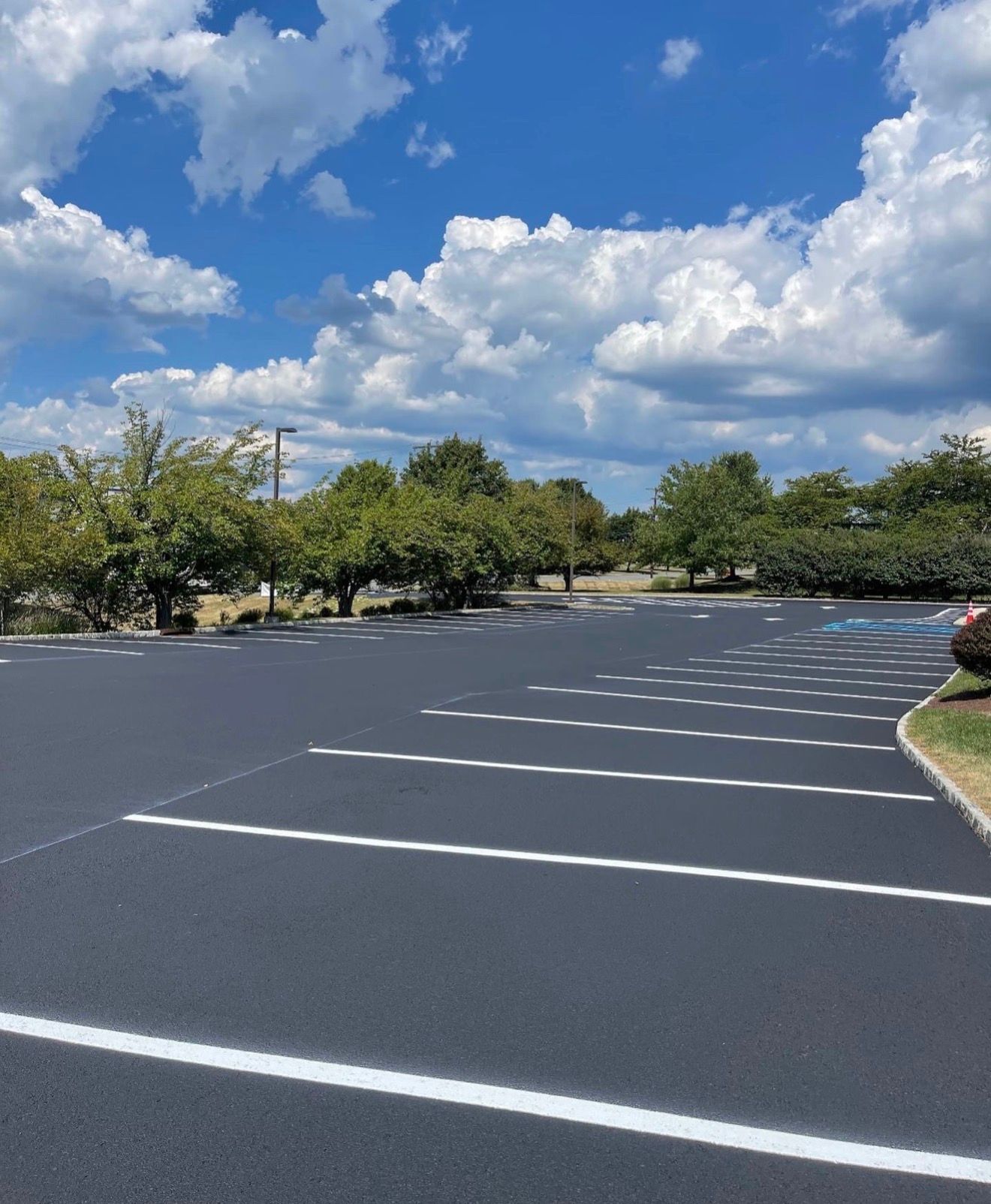 A freshly paved, empty asphalt parking lot with white painted stall lines under a bright, partly cloudy blue sky.