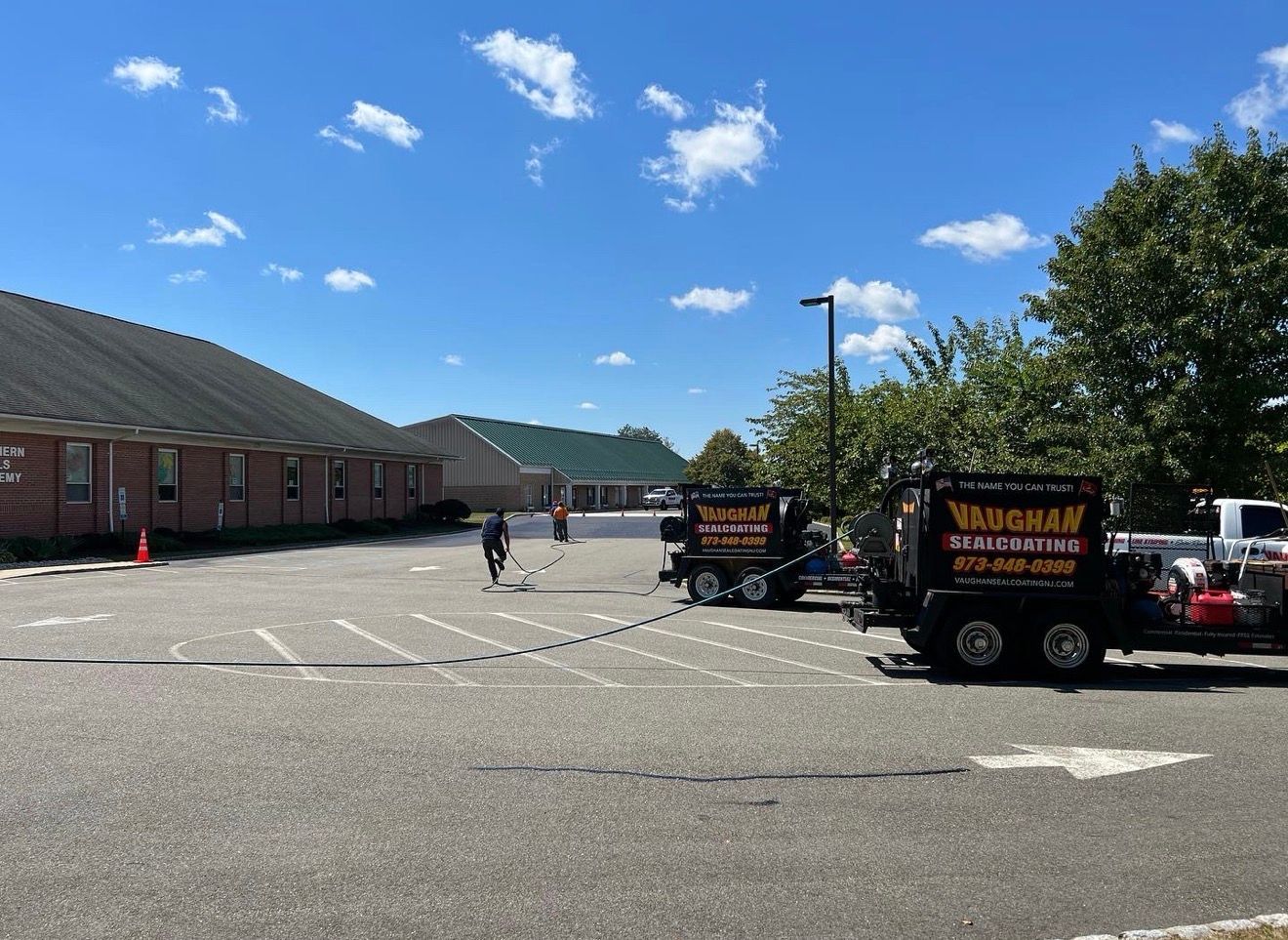A person practices soccer in a parking lot next to a brick building and two parked trailers under a bright blue sky.