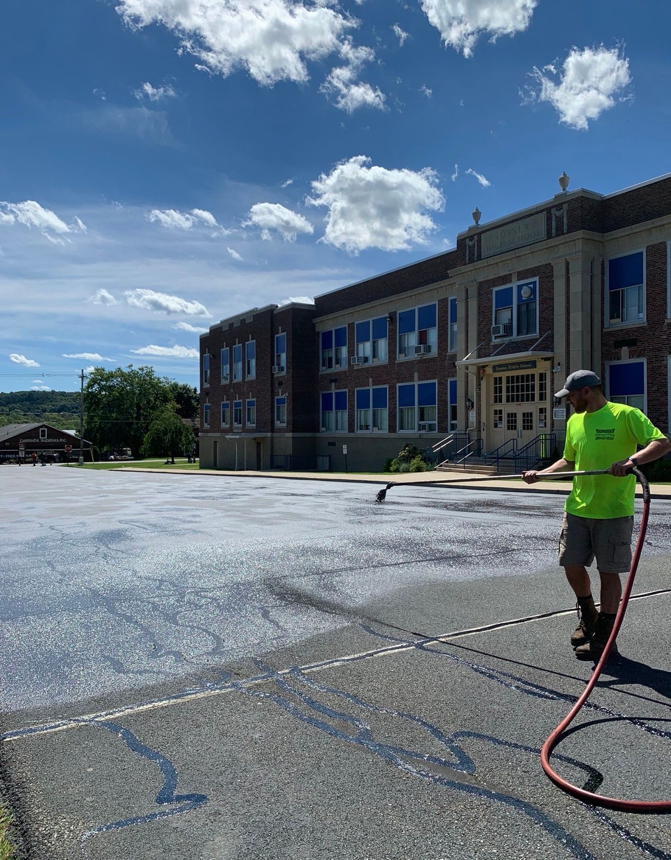 A person in a bright neon shirt uses a pressurized spray hose to apply sealcoating to a paved parking lot at a school.