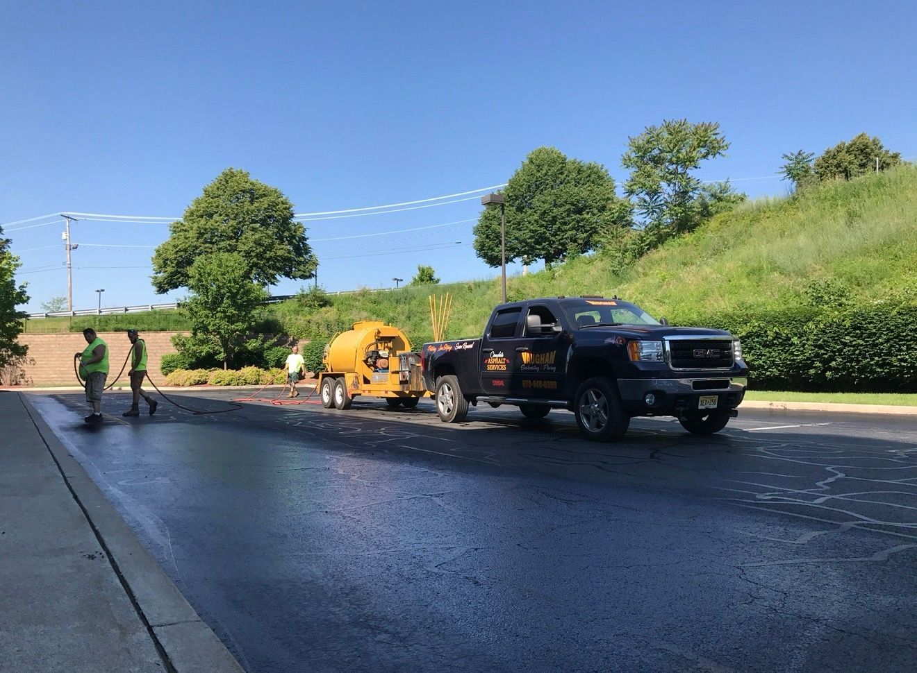 A dark pickup truck towing a yellow sealant tank on a freshly paved black asphalt driveway near a grassy hill.