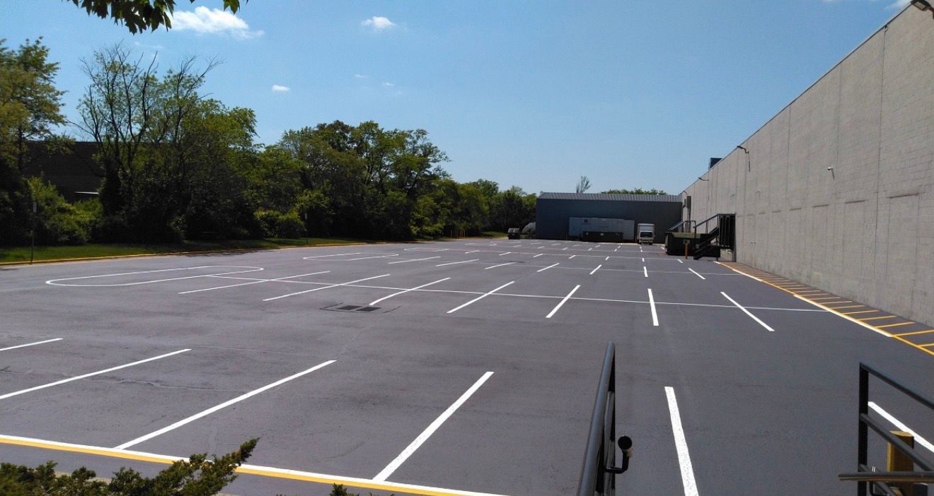 A freshly paved, empty asphalt parking lot with painted white parking lines, bordered by a long wall and green trees.