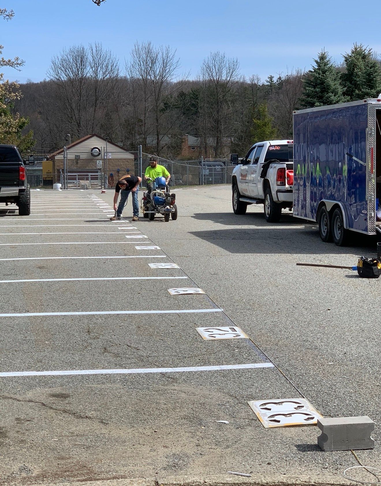 Two workers use a machine to paint white parking space lines and numbers onto a gravel lot next to a utility trailer.