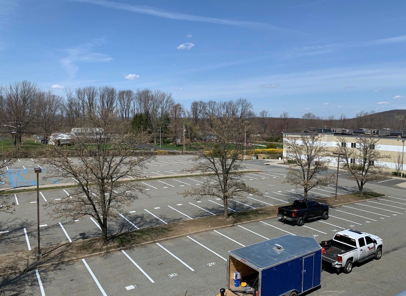 A partially empty parking lot on a sunny day with trees, a black truck, a white pickup, and a blue trailer.