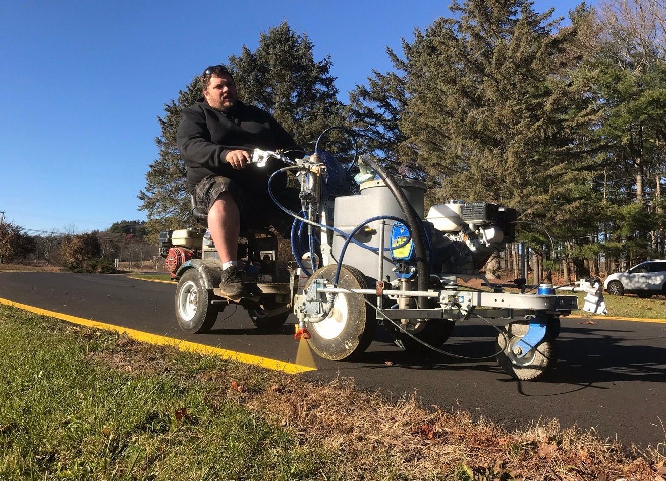 A person on a ride-on machine paints a straight yellow line on an asphalt road in a wooded setting.