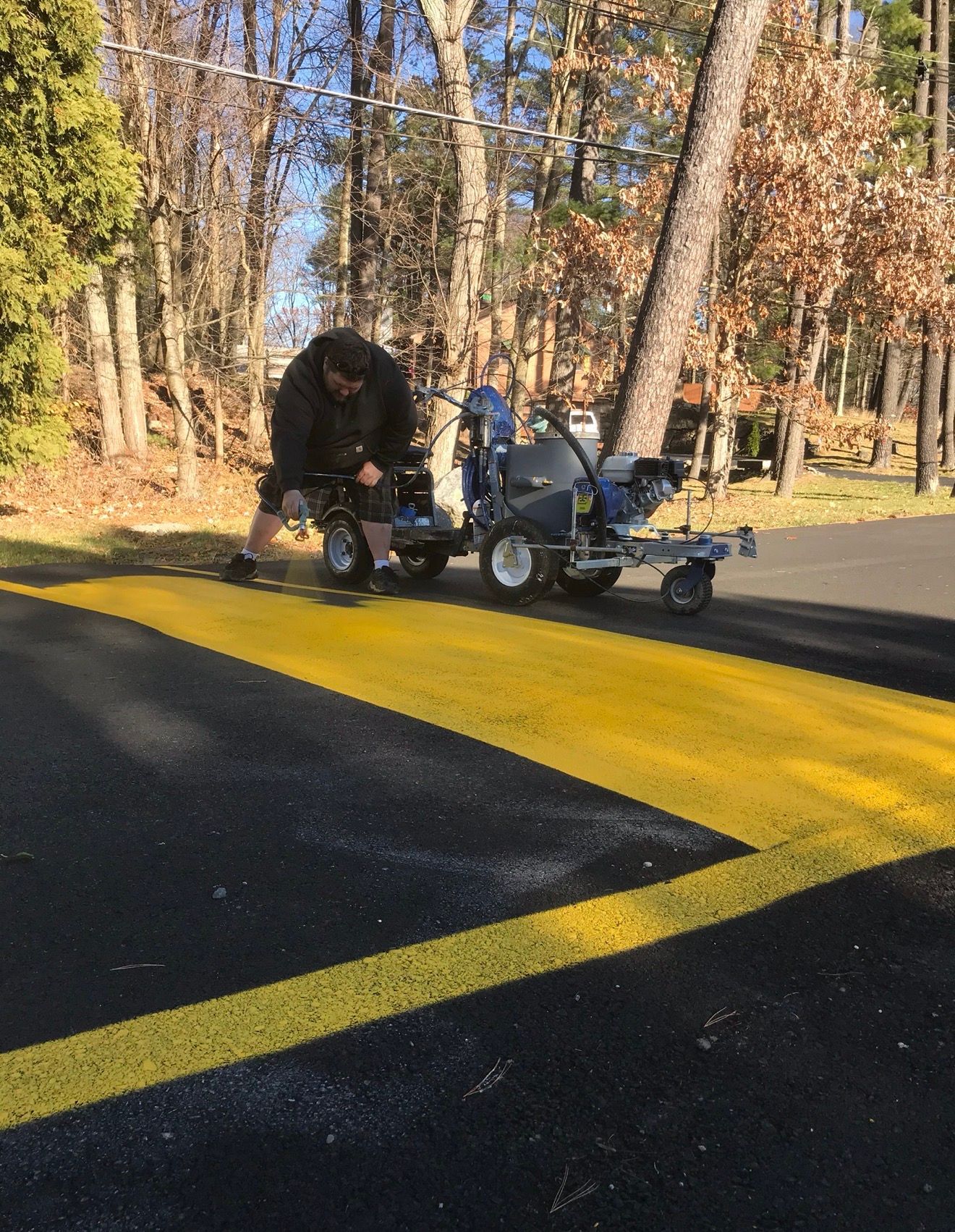 A worker uses a motorized machine to paint yellow lines on fresh black asphalt in a wooded setting.
