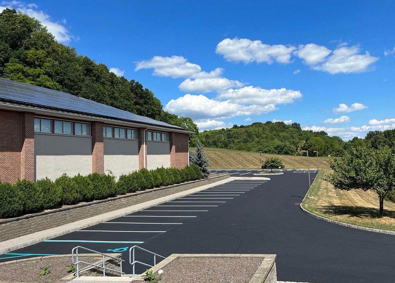 A freshly paved asphalt parking lot next to a brick building, lined with green bushes under a sunny, blue, cloudy sky.